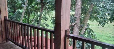 A wooden balcony with a metal railing overlooking a lush garden with various trees and a small pond.