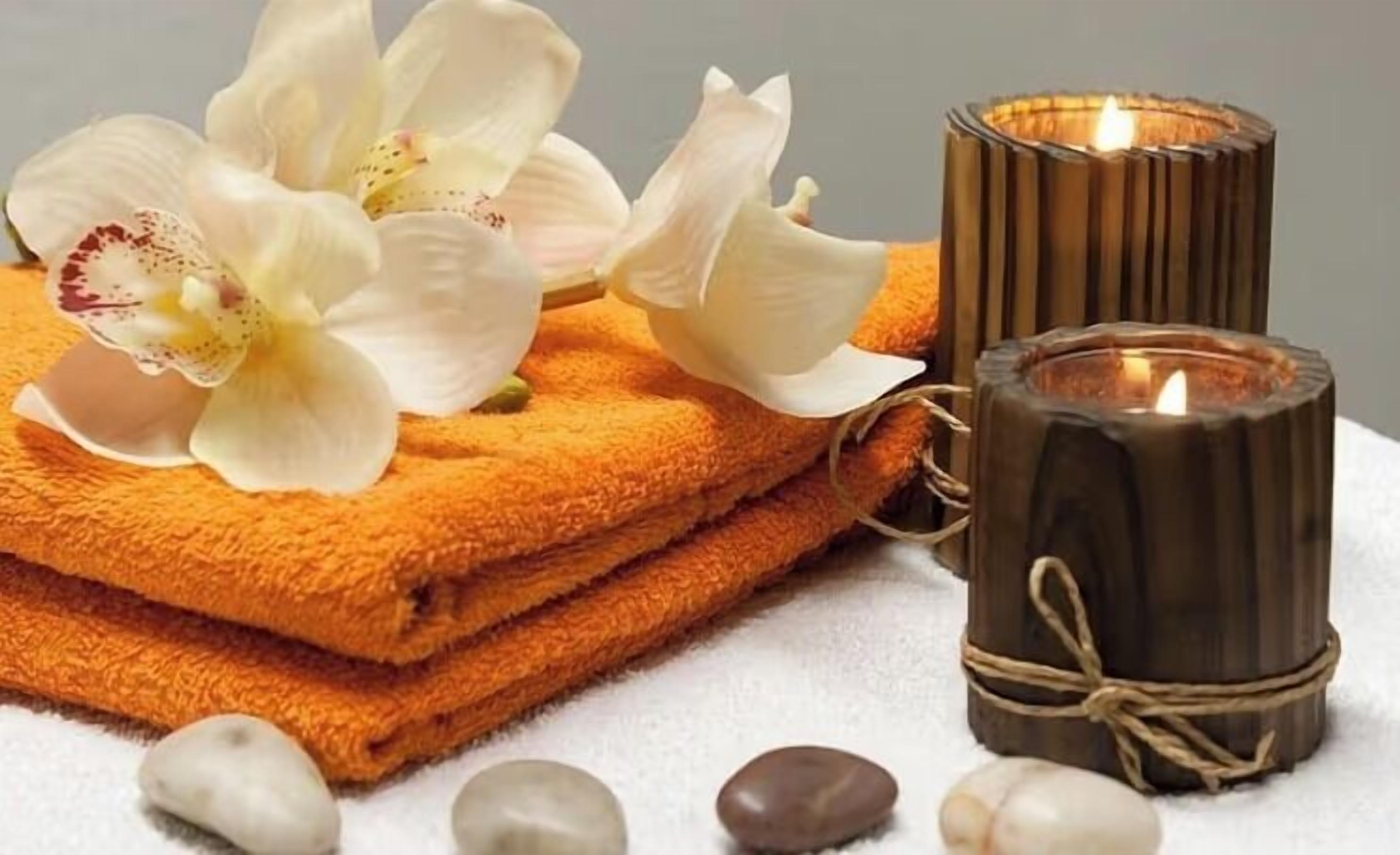 A stack of orange towels, a lit candle, and a white orchid.