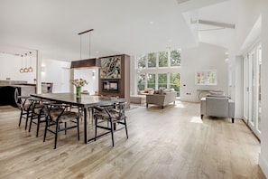 Un vaste espace de vie comprenant une grande table à manger en bois, des chaises modernes et une cheminée. On aperçoit un canapé et un fauteuil au fond de la pièce, et de grandes fenêtres laissent entrer la lumière naturelle.
