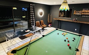 A pool table with a billiards setup, a bar area with glasses, and a view of an outdoor pool table.