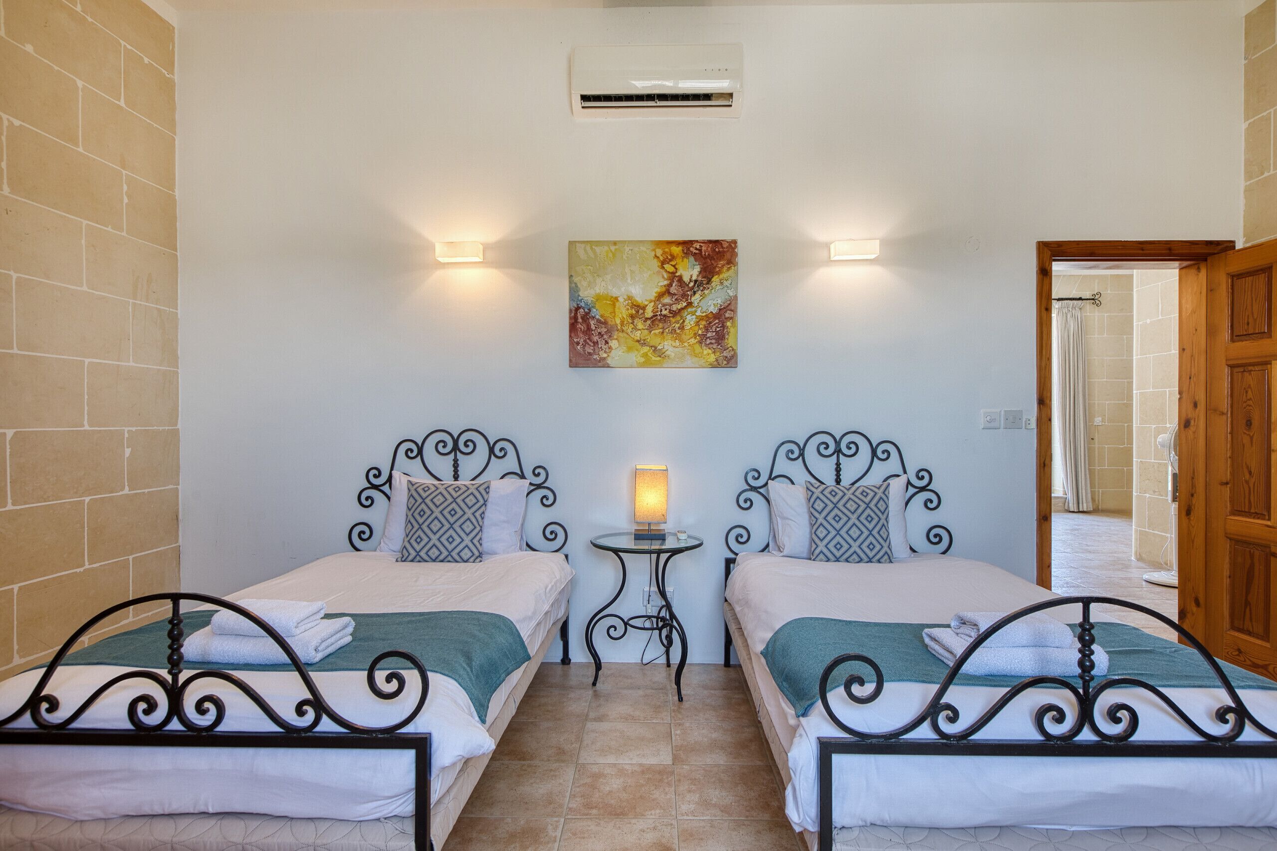 A room with two beds, a small table with a lamp, a painting on the wall, and a wooden door.
