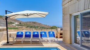 A poolside area with lounge chairs, a canopy, and a view of a hilly landscape. - Rebekah – Ghasri Holiday Home (L-Għasri)