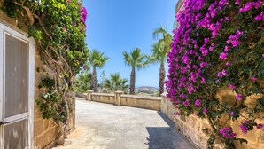A narrow stone pathway flanked by walls covered in purple bougainvillea flowers, leading to a view of palm trees and a distant landscape. - Gharilma IV – Santa Lucija Holiday Home (Ta' Kerċem)