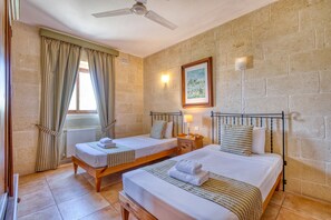 A room with two beds, a window with curtains, a ceiling fan, a wall-mounted light, and a framed picture.