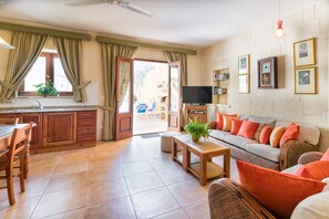 A spacious living area with a sofa, coffee table, and dining table. There is a television, a bookshelf, and a window with curtains.