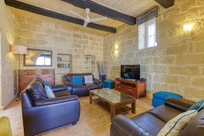 A stone-walled living room with leather sofas, a glass coffee table, a wooden TV stand, and a ceiling fan.