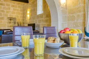 Une table mise pour le petit-déjeuner, avec un mur en pierre en arrière-plan, un saladier rempli de fruits et deux verres de jus d’orange.