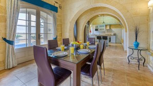 A dining area with a glass table set for four, surrounded by stone walls and an arched doorway leading to a kitchen. - Girardu - Victoria Holiday Home (Victoria)