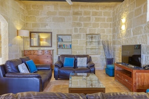 Un salon aux murs de pierre, avec des canapés en cuir, une table basse, un meuble en bois, un téléviseur et des étagères à livres.