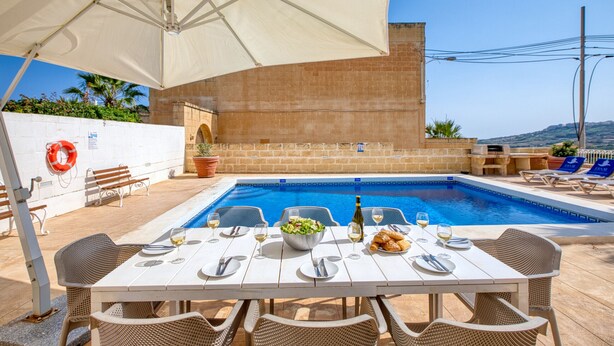 A poolside dining area with a table set for two, a white umbrella, and a view of the pool and landscape. - Hgieri - Ghasri Holiday Home (L-Għasri)