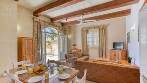 A dining area with a glass table, white chairs, and a breakfast setting. There is a sofa and a television in the living area. - Villagg tal-Fanal - 2BDR (L-Ghasri)