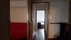 Un pasillo estrecho con un sofá rojo a la izquierda, suelo de baldosas y una cama al fondo.