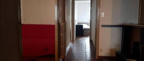 A narrow hallway with a red couch on the left, a tiled floor, and a bed in the background.