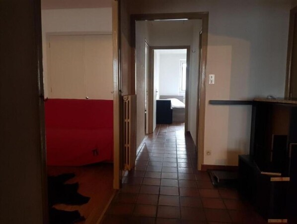 A narrow hallway with a red couch on the left, a tiled floor, and a bed in the background.