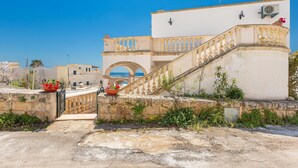 Ein Küstengebäude mit einer Treppe, die zu einem Balkon führt, einer Steinmauer und einem strahlend blauen Himmel.