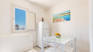 Ein kleines, sauberes Zimmer mit Fenster, Kühlschrank, Tisch mit Stühlen und einem Bild an der Wand.