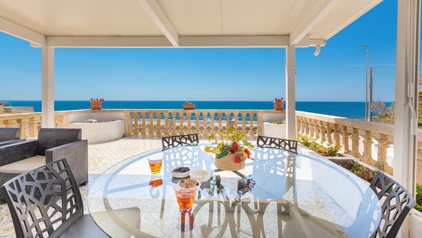 Ein runder Glastisch mit Stühlen, einem Getränk und einer Blumenvase auf einem Balkon mit Blick auf das Meer.