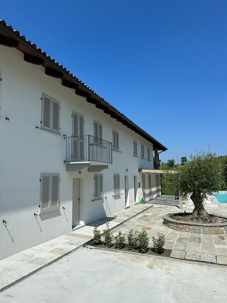 Un bâtiment à deux étages avec une façade blanche, de nombreuses fenêtres dotées de volets, un balcon au deuxième étage et une piscine en arrière-plan.