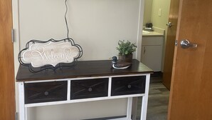 Una habitación con un televisor montado en la pared, un aparador de madera con cajones, un letrero de bienvenida y una planta en maceta.