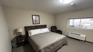 Habitación de hotel con cama, una silla, mesita de noche con lámpara, radiador y ventana con persianas.