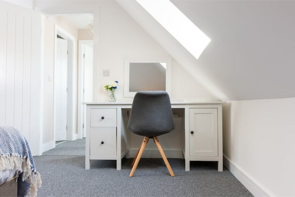 Une chambre avec un plafond en pente, un bureau avec une chaise, un lit et une fenêtre laissant entrer la lumière naturelle.