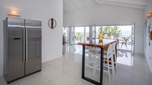 Eine moderne Küche mit einem Edelstahl-Kühlschrank, einem Holztisch mit weißen Stühlen und Blick ins Freie durch große Glasüren.