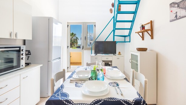Cocina compacta con armarios blancos, microondas y refrigerador. Una mesa de comedor está dispuesta con platos, vasos y cubiertos. Una escalera azul está fijada a la pared y hay un televisor sobre un mueble.
