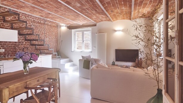 A modern living space with a brick ceiling, a dining area with a wooden table and chairs, a sofa, a TV, and a window with blinds. - AQVAE Unconventional Country House (Piemonte)