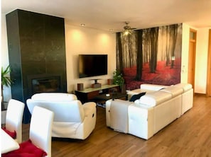 A modern living room with a sectional sofa, a flat-screen TV, and a large forest-themed wall mural. - Bali zen Moderno (Jarana)