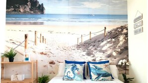 Une chambre avec une fresque représentant une plage, une étagère en bois ornée d’une plante et un lit avec des coussins bleus.