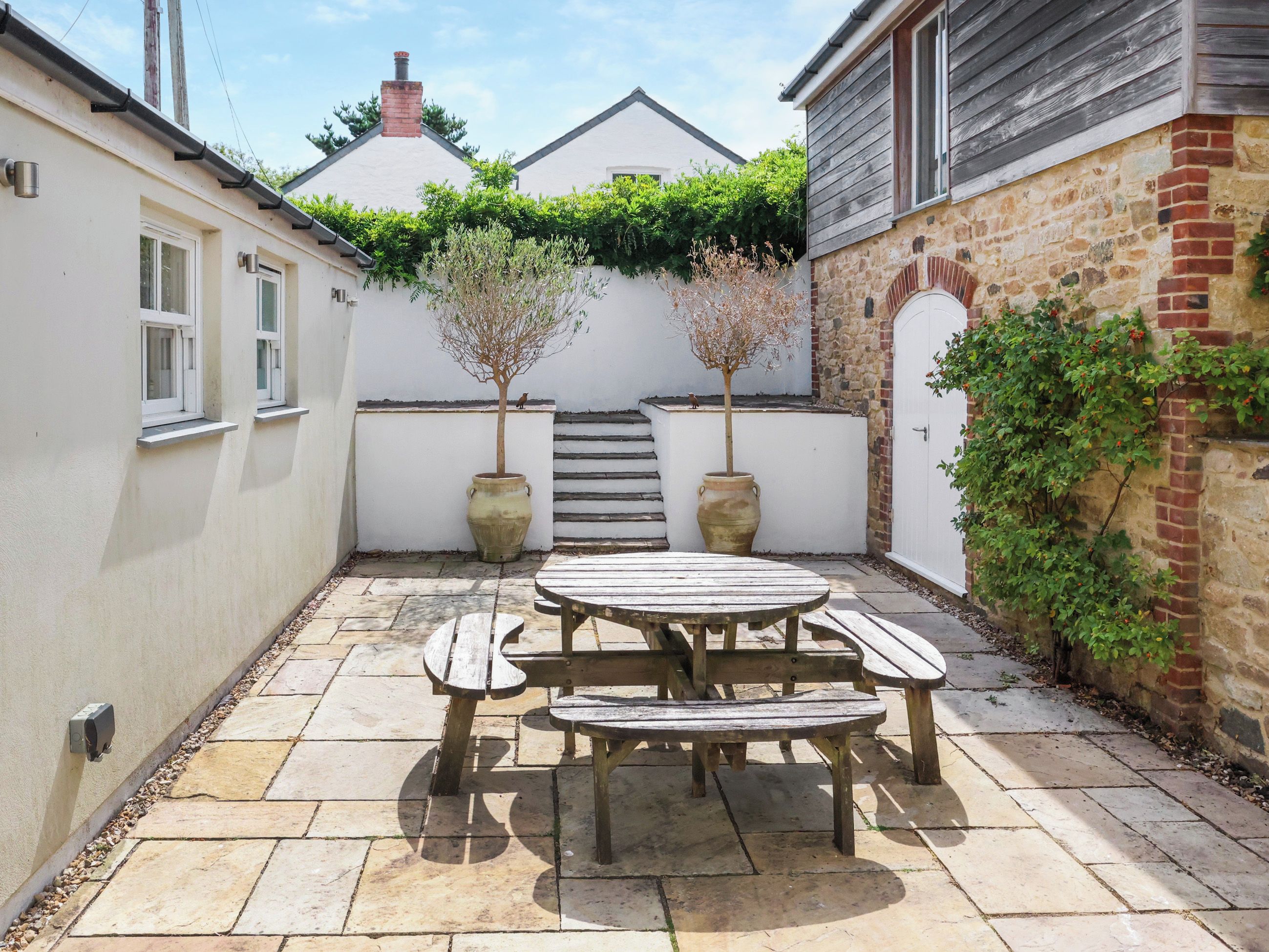 A courtyard with a wooden table and benches, surrounded by walls with windows and a staircase leading to a roof.