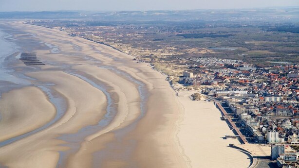 Vista aérea de una ciudad costera con una playa extensa, una zona urbana densa con edificios y un cielo despejado.