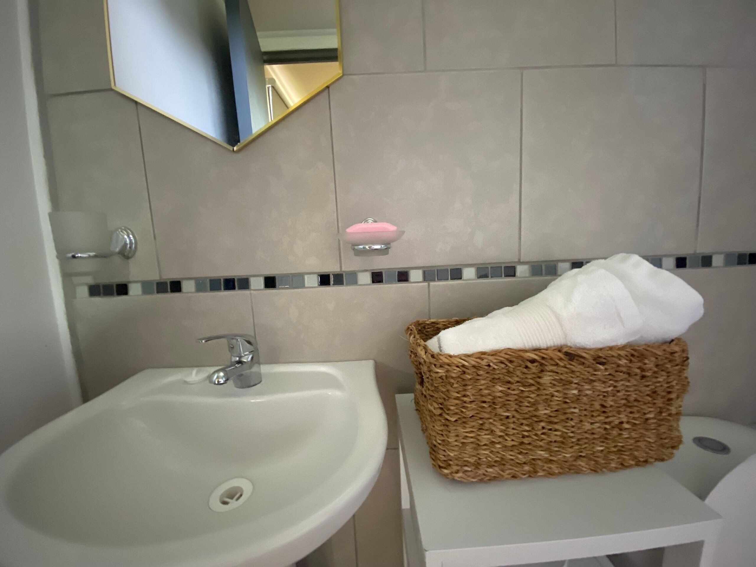 Un baño con un lavamanos blanco, un espejo, un jabonera y una cesta con una toalla.