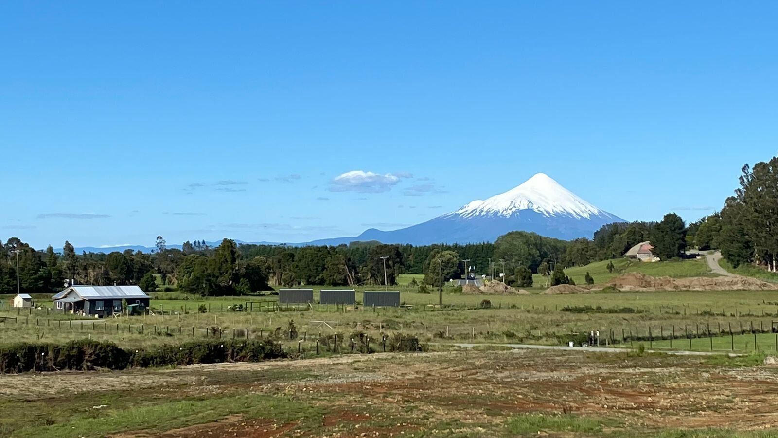A rural landscape with a prominent snow-capped mountain in the background, a fenced field, and a small building with a metal roof.