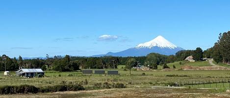 A rural landscape with a prominent snow-capped mountain in the background, a fenced field, and a small building with a metal roof.