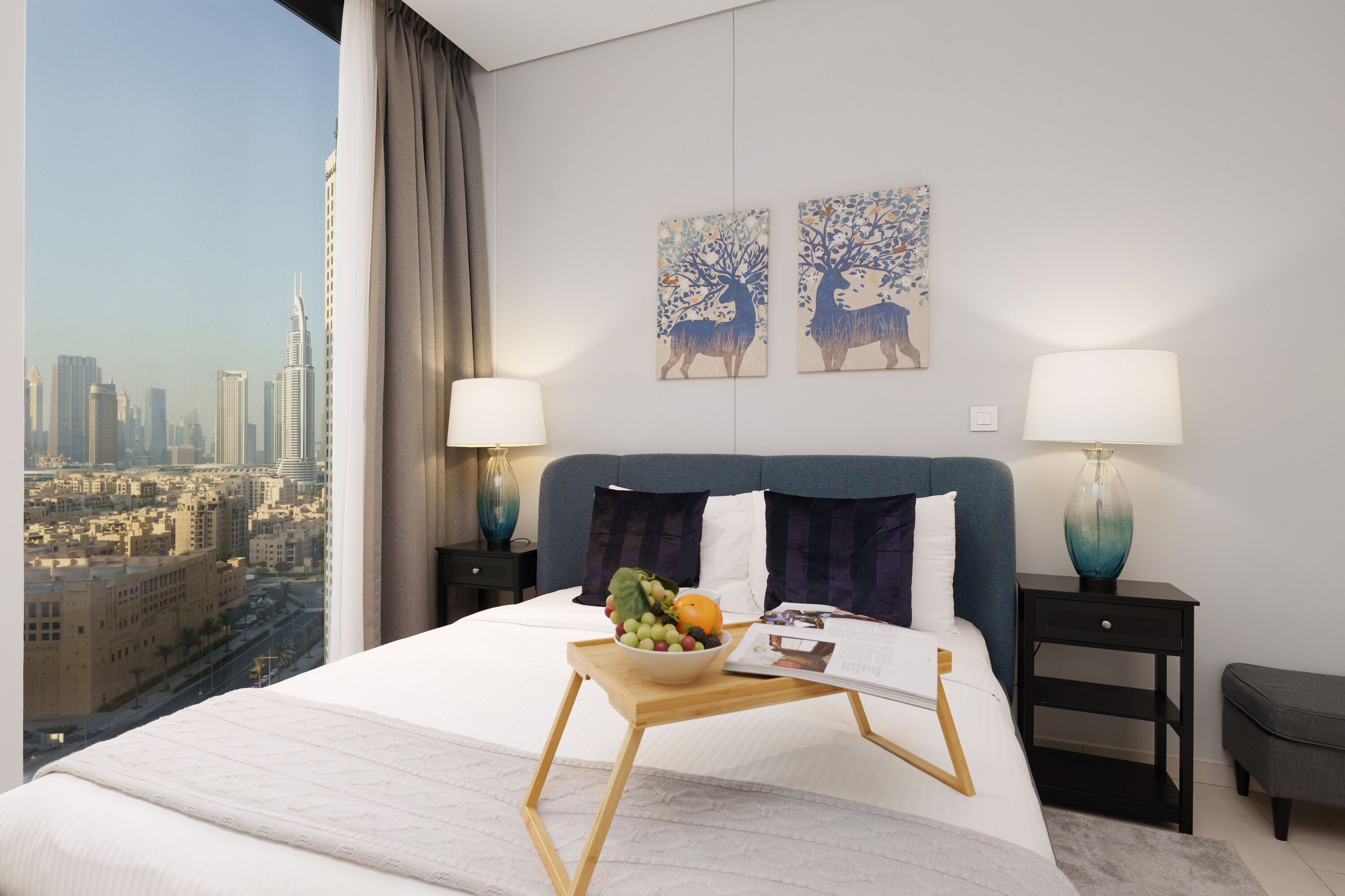 A hotel room with a city view, a bed with a tray table, two side tables with lamps, and artwork on the wall.
