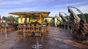 An outdoor seating area with wooden tables and chairs, a central bar area, and a pergola structure.