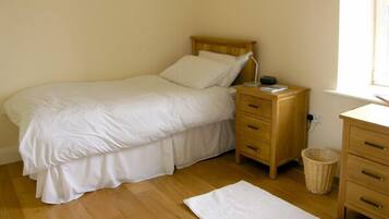 Un dormitorio sencillo y luminoso con una cama de madera, una mesita de noche con lámpara y un pequeño aparador de madera.