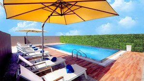 A pool area with lounge chairs and umbrellas, surrounded by a wooden deck and a hedge. - Standard Room In Jaddaf By E R (دبي)