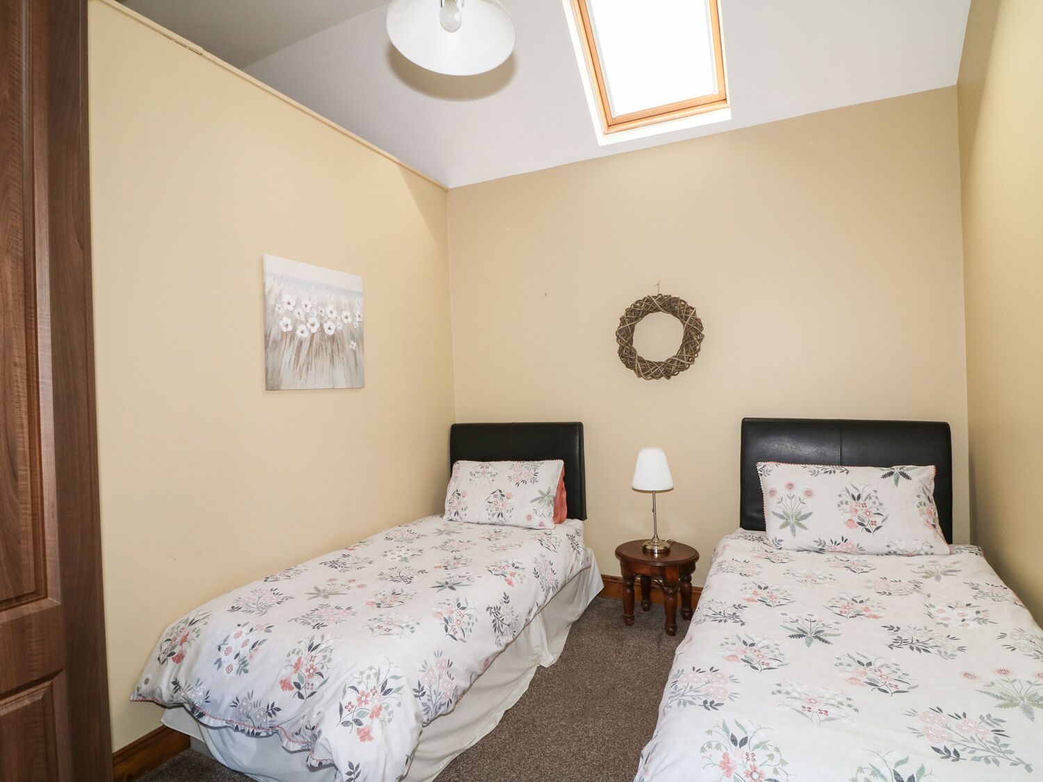A bedroom with two single beds, a small bedside table with a lamp, a wall-mounted wreath, and a ceiling light.
