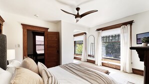 Habitación de hotel con una cama grande, puerta de madera, ventanas con cortinas, un ventilador de techo y un televisor sobre un mueble de madera.
