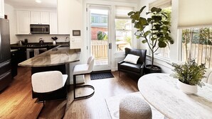 Una cocina moderna con armarios blancos, microondas integrado y una isla grande. Hay una zona de estar con una silla y reposapiés, y una planta en maceta junto a la ventana.