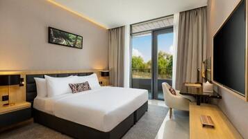 A modern hotel room with a large bed, a desk, a chair, a TV, and a balcony with a view of greenery.