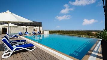 A pool area with blue lounge chairs and umbrellas, a covered seating area, and a view of the landscape.