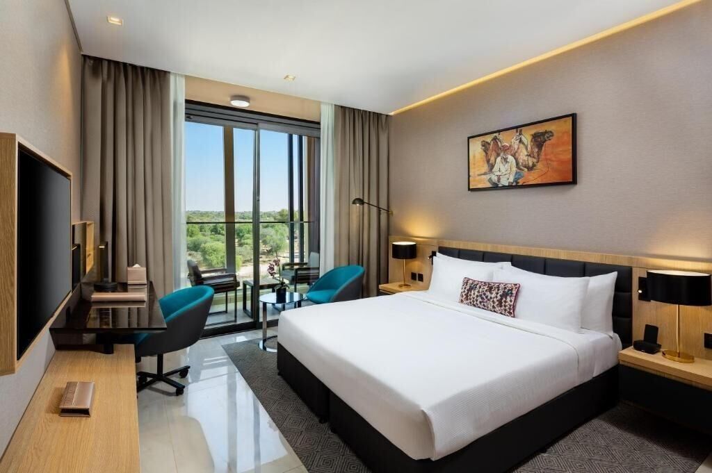 A modern hotel room with a large bed, a desk with a chair, a small table, and a balcony with a view.