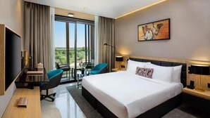 A modern hotel room with a large bed, a desk with a chair, a small table, and a balcony with a view. - Deluxe Room in Mirdif By E R (Dubai)