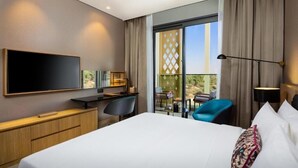 A hotel room with a large bed, a desk, a chair, a TV, and a balcony with a view. - Deluxe Room in Mirdif By E R (Dubai)