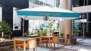 Un espace extérieur aménagé avec des sièges, un panneau indiquant « Café-lounge », des meubles en osier et des plantes en pot.