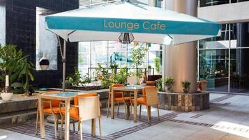 Un espace extérieur aménagé avec des sièges, un panneau indiquant « Café-lounge », des meubles en osier et des plantes en pot.