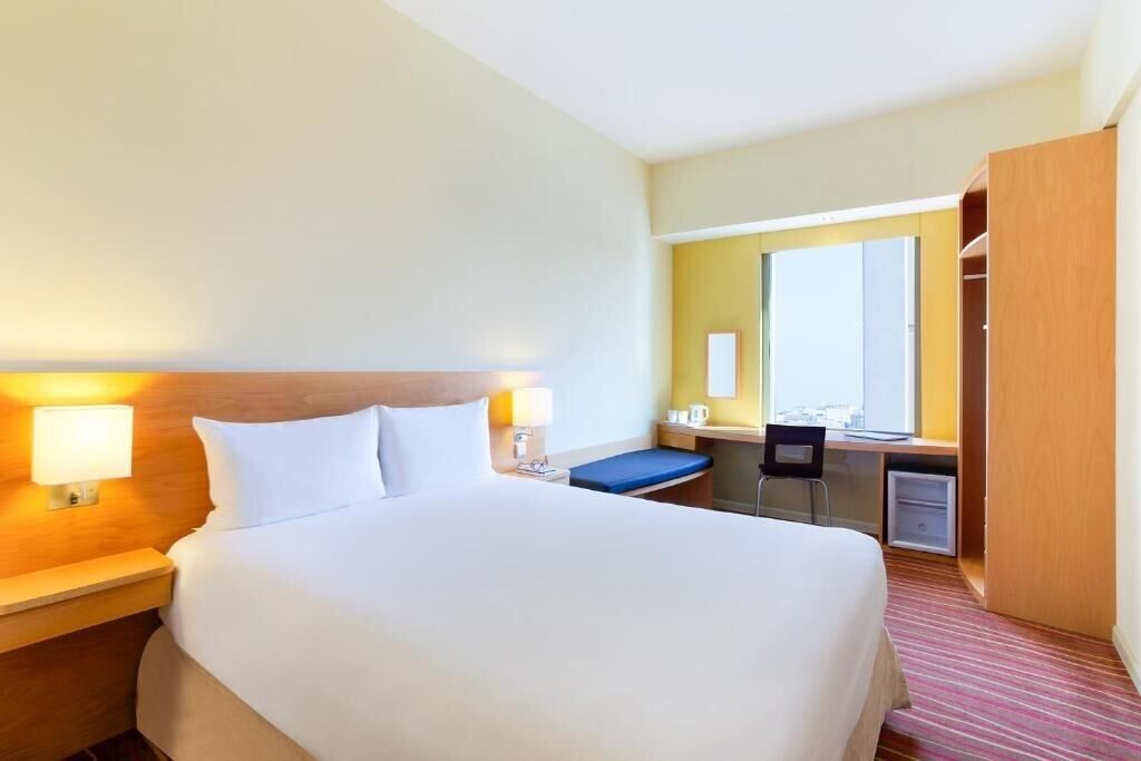Een hotelkamer met een groot bed, nachtlampjes, een bureau met stoel, een spiegel en een kledingkast.